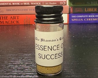 May include: A small glass bottle with a black cap, labeled "Essence of Success." The bottle contains a light brown powder. The background includes books with titles like "Sigil Magick" and "Cunningham's Encyclopedia."