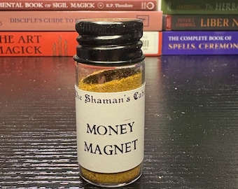 May include: A small glass jar with a black lid, filled with a golden-brown powder. The jar has a white label with the words "MONEY MAGNET" in black text. The background includes a stack of books.