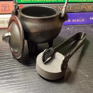 May include: A black cast iron cauldron with three legs, a matching lid, and a metal handle. Beside the cauldron sits a pair of black metal tongs gripping a small, round, gray charcoal disc. The set is designed for burning incense or resins.