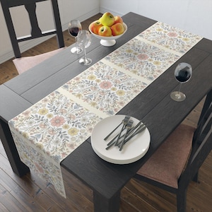 May include: A white table runner with a floral pattern in shades of pink, yellow, and gray. The runner is laid on a dark wood table with a plate and silverware.