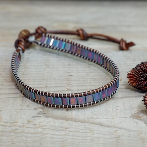 Puede incluir: Una pulsera artesanal con cuentas rectangulares iridiscentes en tonos azules, morados y verdes, cosidas sobre una banda de cuero marrón con hilo blanco. La pulsera tiene un cierre de cuero. Flores secas en el fondo.