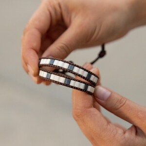 Puede incluir: Una pulsera envolvente con una banda de cuero marrón y cuentas rectangulares blancas y grises. La pulsera se sostiene en las manos de una persona contra un fondo borroso. La pulsera se envuelve dos veces alrededor de la muñeca.