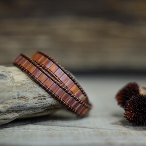 Puede incluir: Un brazalete de doble vuelta hecho con pequeñas cuentas cuadradas iridiscentes de color cobre. El brazalete está sobre una superficie de madera.