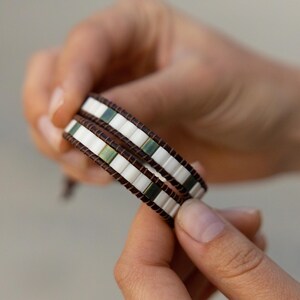 Puede incluir: Dos pulseras envolventes con una banda de cuero marrón y cuentas rectangulares blancas, verdes y marrones. Las pulseras están sujetas en las manos de una persona.