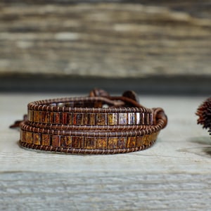May include: A brown leather wrap bracelet with small, square, iridescent beads. The bracelet is wrapped around itself twice and has a leather tie closure.