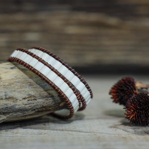 May include: A handcrafted bracelet featuring a repeating pattern of white rectangular beads and brown leather. The bracelet is wrapped around a piece of wood, with dried seed heads in the background. The bracelet's design is simple and elegant.