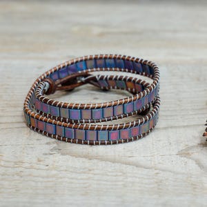 May include: A double wrap bracelet made with iridescent square beads and brown leather. The bracelet is on a wooden surface.