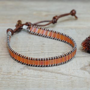 Puede incluir: Una pulsera artesanal con cuentas rectangulares naranjas y blancas, ensartadas en una banda de cuero marrón. La pulsera tiene un cierre de cuero. El diseño está acentuado con costuras blancas. La pulsera mide aproximadamente 18 cm de largo.