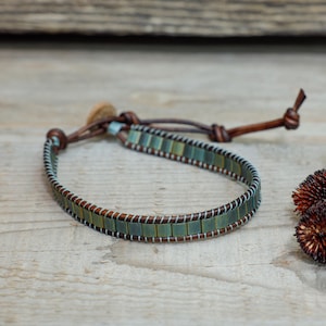 Puede incluir: Una pulsera artesanal con una banda de pequeñas cuentas cuadradas, verde azulado y marrón. Las cuentas están cosidas sobre una banda de cuero marrón con hilo blanco. La pulsera tiene un cierre de cuero.