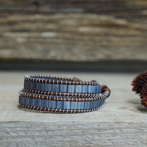 Puede incluir: Una pulsera envolvente con cuentas rectangulares azul-gris y correas de cuero marrón. La pulsera está envuelta sobre sí misma dos veces. Las cuentas están ensartadas con hilo blanco. Flores secas en el fondo.