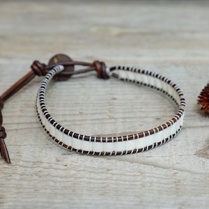 Puede incluir: Una pulsera artesanal con una banda color crema y detalles en marrón oscuro. La pulsera es de cuero y presenta un cierre de botón y una correa de cuero anudada. Flores secas en el fondo.