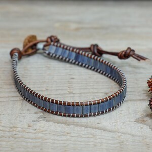 Puede incluir: Una pulsera azul y marrón con un diseño envolvente. La pulsera presenta pequeñas cuentas azules rectangulares, cuero marrón y costuras blancas. La pulsera mide aproximadamente 18 cm de largo.
