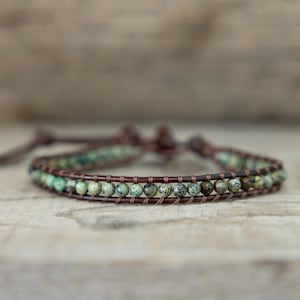 May include: A wrap bracelet featuring small, round, green and brown beads. The beads are strung on a brown leather cord, with a second leather cord used for wrapping. The bracelet is displayed on a light brown wooden surface.