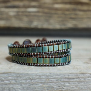 Puede incluir: Un brazalete de doble vuelta con cuentas cuadradas en tonos verdes y marrones. El brazalete está hecho de cuero y tiene un hilo blanco que atraviesa las cuentas.