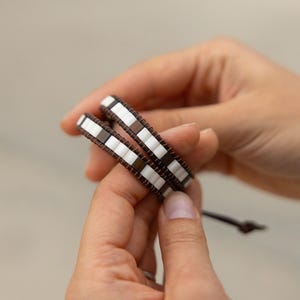 Puede incluir: Dos pulseras envolventes con una banda de cuero marrón y un patrón de cuentas rectangulares blancas y marrones. Las pulseras están en las manos de una persona.