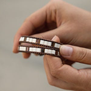 May include: Two wrap bracelets with white and brown rectangular beads. The bracelets are wrapped around each other. The bracelets are held by a person's hands.