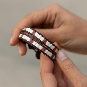 Puede incluir: Una pulsera envolvente con una banda de cuero marrón y un patrón de cuentas blancas y burdeos. La pulsera se sostiene en las manos de una persona, mostrando su diseño. La pulsera se envuelve alrededor de la muñeca varias veces.