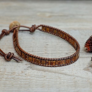 Puede incluir: Una pulsera de cuero marrón con cuentas cuadradas texturizadas. La pulsera es ajustable con un cordón de cuero y un cierre de cuentas de madera. El fondo incluye flores secas sobre una superficie de madera.