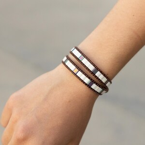 Puede incluir: Dos pulseras envolventes con cuentas rectangulares blancas y detalles iridiscentes envuelven una muñeca. Las pulseras tienen una banda de cuero marrón. El fondo está borroso.