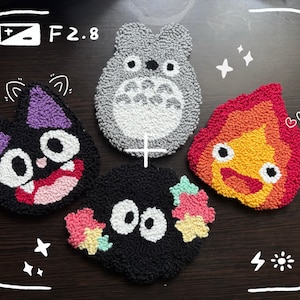 Può includere: Quattro sottobicchieri colorati e fatti a mano con personaggi dei cartoni animati. I sottobicchieri includono un Totoro grigio, un gatto nero con orecchie viola, uno sprite di fuliggine nero e un demone di fuoco rosso e giallo. L'immagine è incorniciata con un bordo simile a una fotocamera.