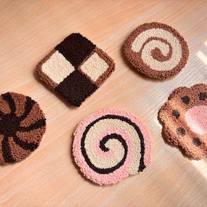 May include: Five handmade, tufted coasters in various shapes and designs. Includes a checkerboard square, a swirl, a spiral, and a flower-shaped coaster. The coasters feature brown, beige, and pink colours.
