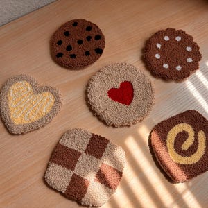 May include: Six handmade, tufted coasters in various shapes and designs. The coasters feature cookie-inspired patterns, including chocolate chips, a heart, and a checkerboard. The colour palette includes shades of brown, beige, and yellow.