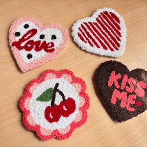May include: Four handmade heart-shaped coasters. One pink coaster has the word "love" in red. Another has red stripes. A third has cherries, and the last has "Kiss Me" in pink.