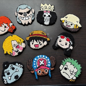 Puede incluir: Una colección de diez posavasos de punch needle con formas variadas que presentan rostros de personajes de dibujos animados, incluyendo figuras con cabello naranja, una calavera con corona, un sombrero de paja y un sombrero azul con cruz rosa.