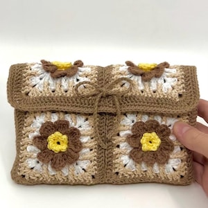 Könnte beinhalten: Handgefertigte Häkeltasche in Braun-, Beige- und Weißtönen mit floralem Design. Die Tasche hat einen passenden Kordelverschluss. Die Blumen haben gelbe Zentren. Das Gesamtdesign ist ein quadratisches Muster.