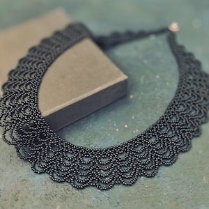 Może przedstawiać: Czarny naszyjnik z koralików z falistym brzegiem. Naszyjnik jest prezentowany na szarej skrzynce na turkusowym tle. Złożona praca z koralików tworzy delikatny, teksturowany wygląd, odpowiedni na specjalne okazje.