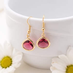 May include: A pair of gold-colored drop earrings with teardrop-shaped, ruby-red gemstones. The earrings are displayed against a white background with white flowers. The gemstones are set in a gold bezel.