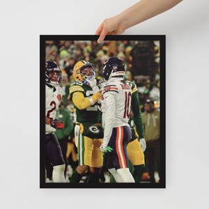 Op de afbeelding: Ingelijste print met een footballwedstrijd. Twee spelers in groen-gele uniformen staan tegenover een speler in een wit en marineblauw uniform. De spelers dragen helmen en bescherming. De achtergrond toont een menigte toeschouwers.