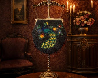 Puede incluir: Un bolso negro vintage con cuentas y un diseño de pavo real en azul, verde, amarillo y naranja. Se exhibe sobre un soporte dorado decorativo. El fondo presenta un interior oscuro y ornamentado con una pintura y muebles antiguos.