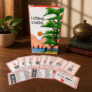 May include: A deck of I Ching Cards with a box featuring a landscape illustration with a tree, sun, and mountains. The cards are white with red borders and Chinese characters. A decorative incense burner and books are also visible.