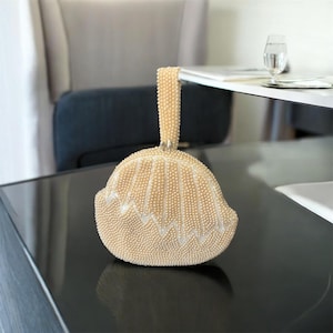 Puede incluir: Un bolso de mano vintage con cuentas de color crema con asa. El bolso tiene forma redondeada con borde festoneado y un patrón en zigzag. El asa también está adornada con cuentas. La bolsa está sobre una superficie negra.