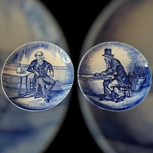 May include: Two antique blue and white porcelain plates. Each plate features a detailed illustration of a man in a seated position. One plate shows a man with a pipe, the other shows a man with a hat. The plates are likely decorative.