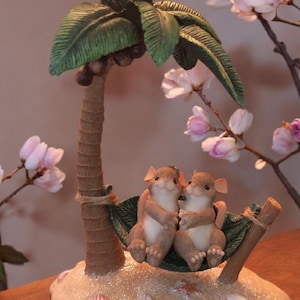 Può includere: Una statuina stravagante con due topi che si rilassano in un'amaca sotto una palma in miniatura. La base è progettata per assomigliare a una spiaggia sabbiosa, completa di conchiglie. La palma ha foglie verdi e noci di cocco.