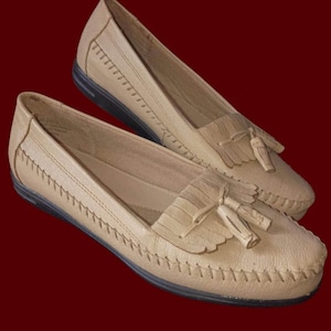 May include: A pair of beige leather loafers with decorative stitching and tassel accents. The shoes have a rounded toe and a low heel, suitable for casual wear. The soles are black.