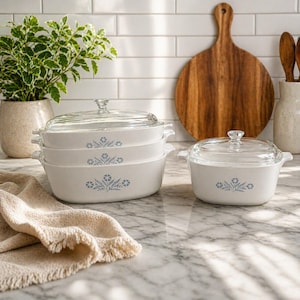 May include: A collection of white ceramic casserole dishes with clear glass lids. Each dish has a blue floral pattern. The dishes are stacked on a marble surface, with a wooden chopping board and a plant in the background.