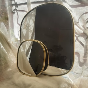 Puede incluir: Dos espejos decorativos con marcos dorados. Uno es rectangular con esquinas redondeadas, el otro más pequeño y ovalado. Los espejos están colocados frente a una cortina blanca transparente.