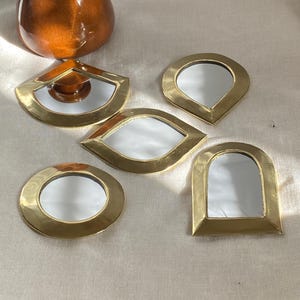 May include: A collection of five small, decorative mirrors in various shapes: a circle, a teardrop, an eye, a triangle, and a rounded rectangle. Each mirror has a gold-colored metal frame. A brown ceramic vase is in the background.