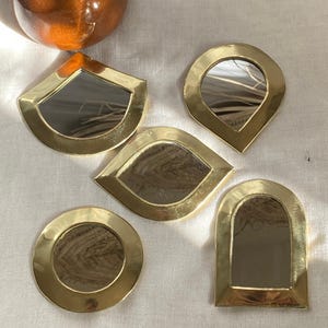 Può includere: Cinque specchi decorativi in ottone di varie forme: un cerchio, una goccia, una foglia, un quarto di cerchio e un arco. Ogni specchio ha una superficie riflettente e una cornice in ottone lucido. Un vaso di ceramica marrone è sullo sfondo.