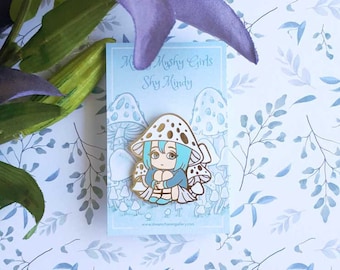 May include: A gold-toned enamel pin featuring a cartoon girl with blue hair and a white dress sitting on a mushroom. The mushroom is white with black spots. The pin is on a white card with blue and green floral designs. The card has the text "Mushy Girls Sky Mindy" and a website address.