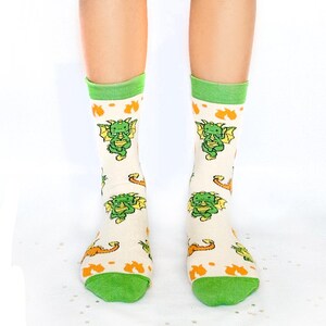 May include: Cream-coloured socks with green and orange accents. The socks feature a repeating pattern of green cartoon dragons with yellow wings. The top and toe of the socks are green.