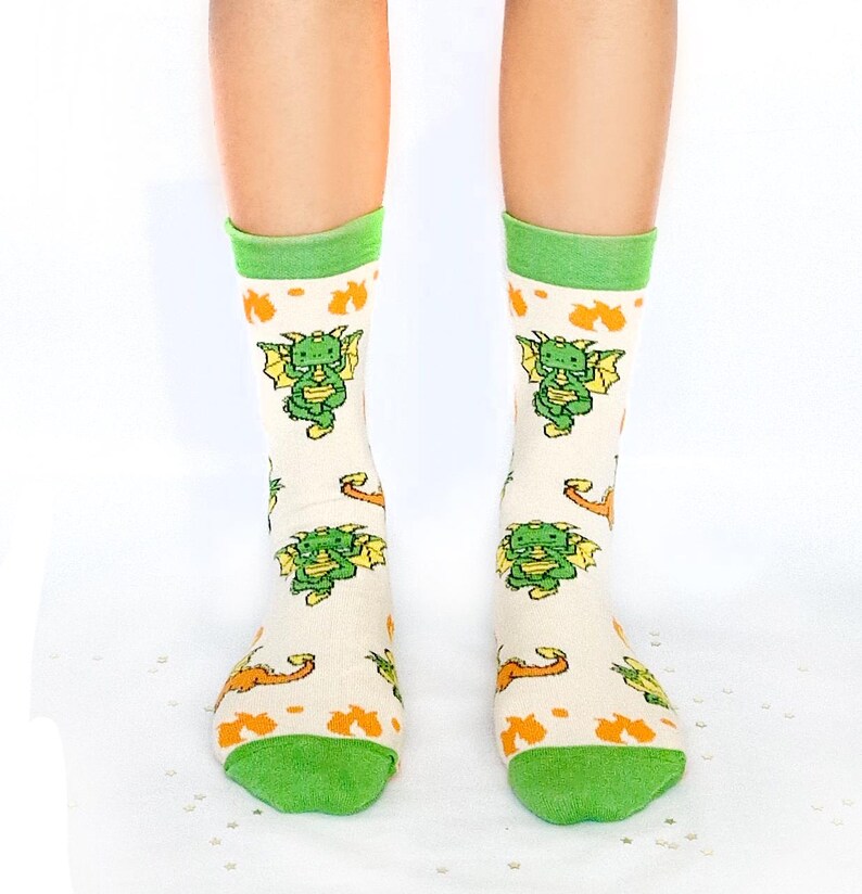 May include: Cream-coloured socks with green and orange accents. The socks feature a repeating pattern of green cartoon dragons with yellow wings. The top and toe of the socks are green.