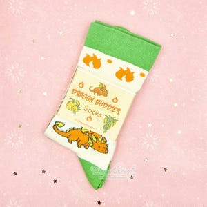 May include: A pair of green and white socks with orange dragon graphics. The socks feature the text "Dragon Buddies Socks" on the packaging. The socks are folded and displayed on a pink background with star and sunburst patterns.