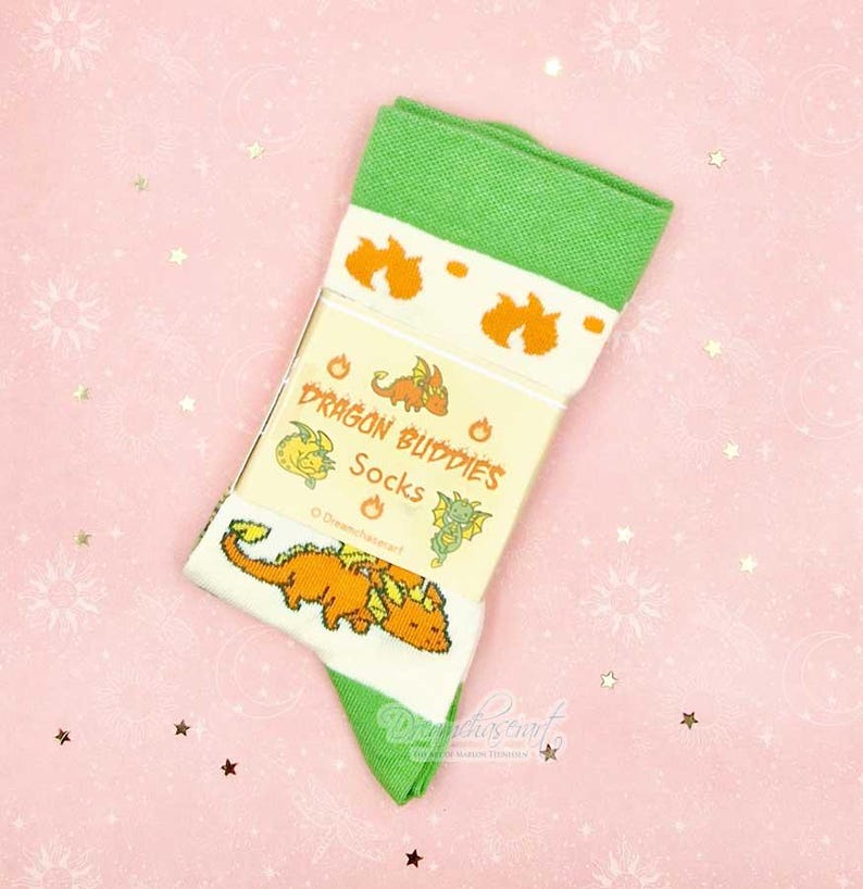 May include: A pair of green and white socks with orange dragon graphics. The socks feature the text "Dragon Buddies Socks" on the packaging. The socks are folded and displayed on a pink background with star and sunburst patterns.