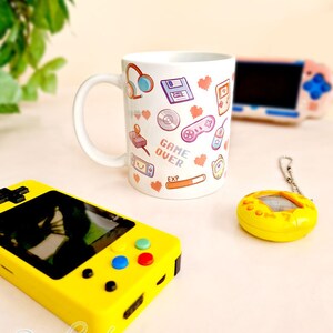 Pu&ograve; includere: Una tazza in ceramica bianca con un design colorato che presenta icone di videogiochi retr&ograve;, cuori e il testo "GAME OVER". La tazza &egrave; circondata da una console di gioco portatile gialla, un giocattolo digitale per animali domestici giallo e una console di gioco portatile rosa.