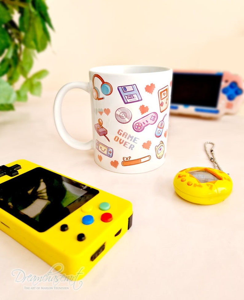 Pu&ograve; includere: Una tazza in ceramica bianca con un design colorato che presenta icone di videogiochi retr&ograve;, cuori e il testo "GAME OVER". La tazza &egrave; circondata da una console di gioco portatile gialla, un giocattolo digitale per animali domestici giallo e una console di gioco portatile rosa.
