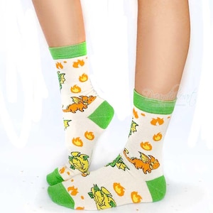 May include: Cream socks with green cuffs, heels, and toes. The socks display a repeating pattern of orange dragons and yellow flames. The socks are knee-high and made of a soft material.